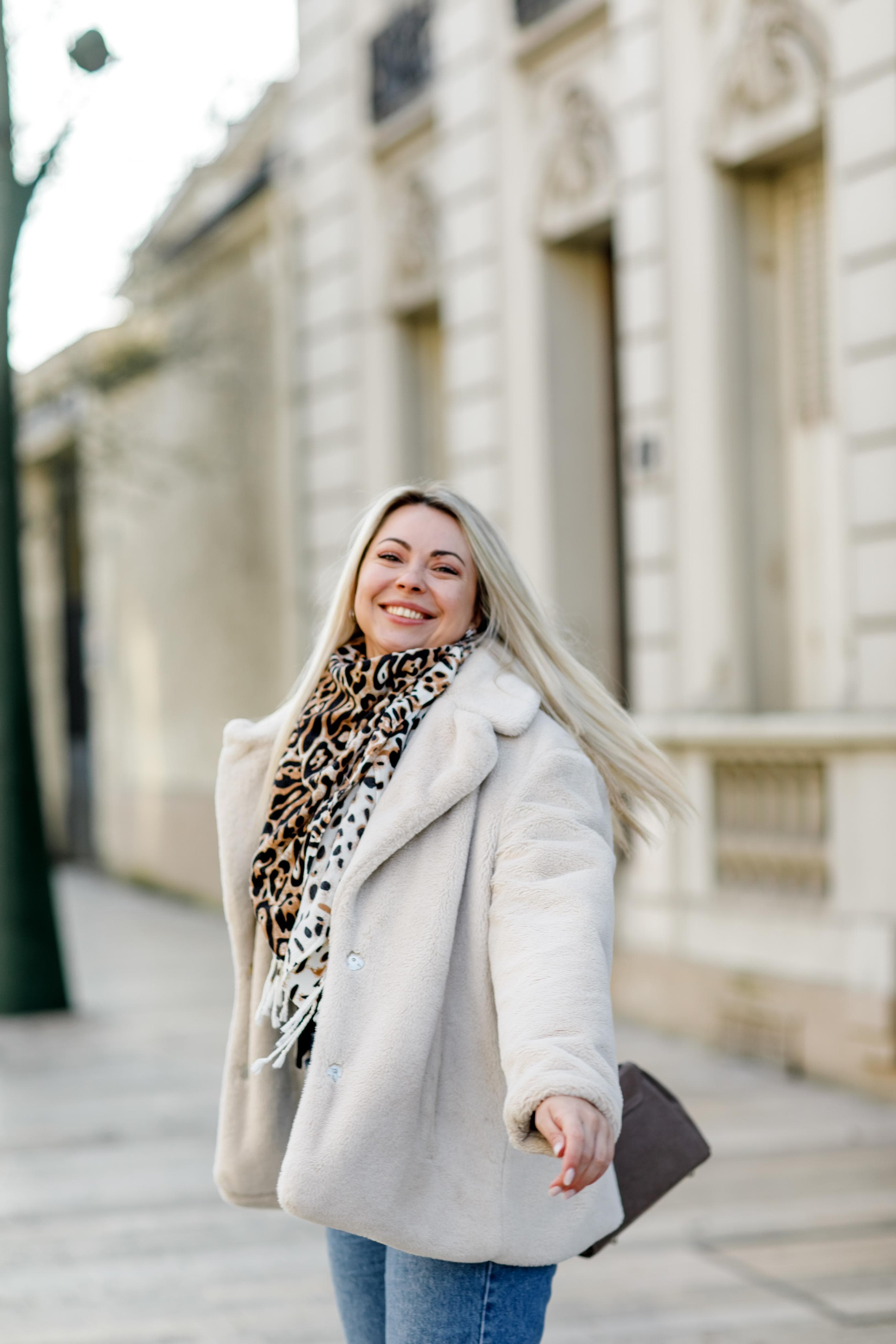 Femme blonde souriante portant un manteau blanc et une écharpe léopard marchant en ville.