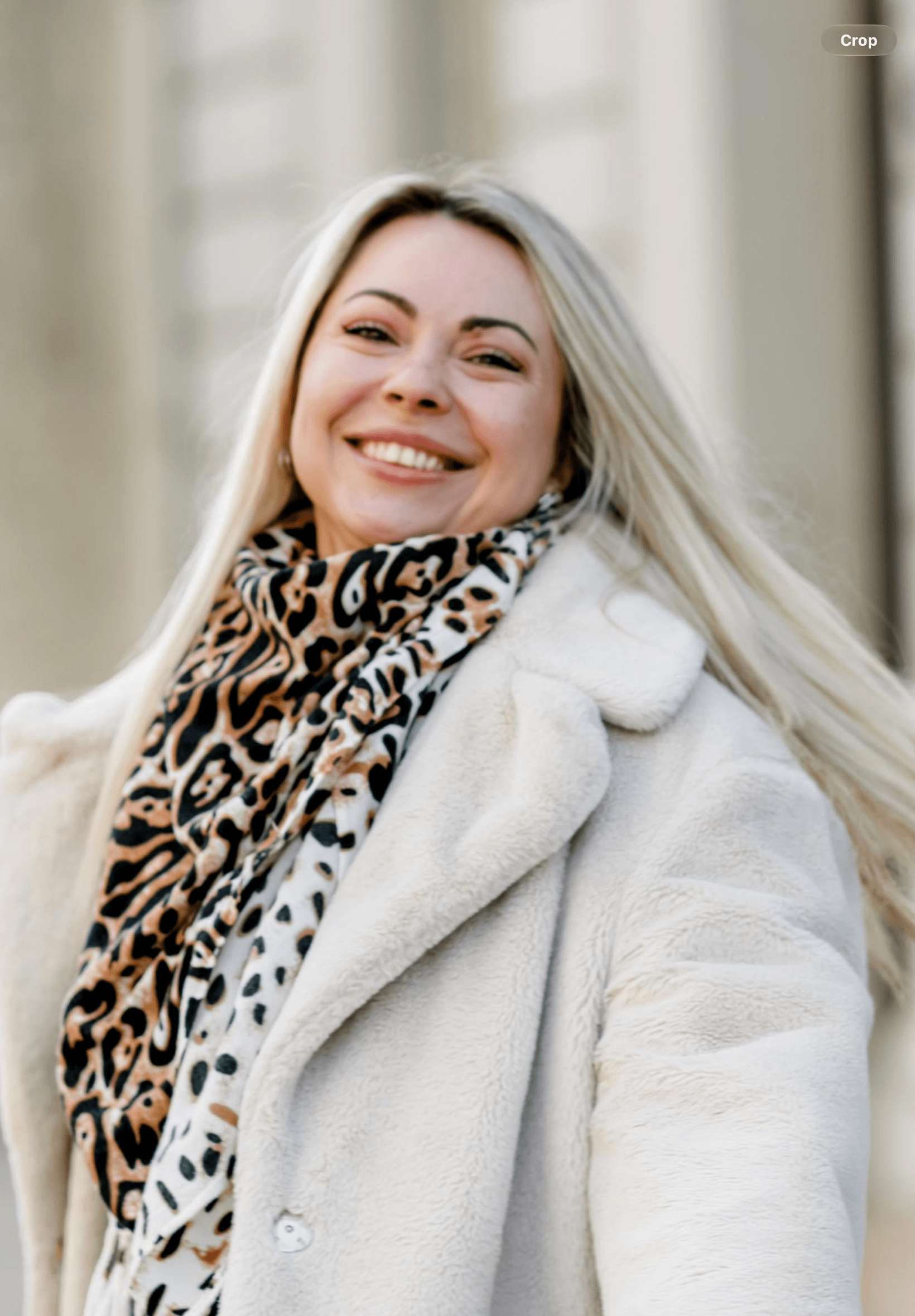 Femme blonde souriante portant un manteau crème en fausse fourrure et une écharpe léopard.