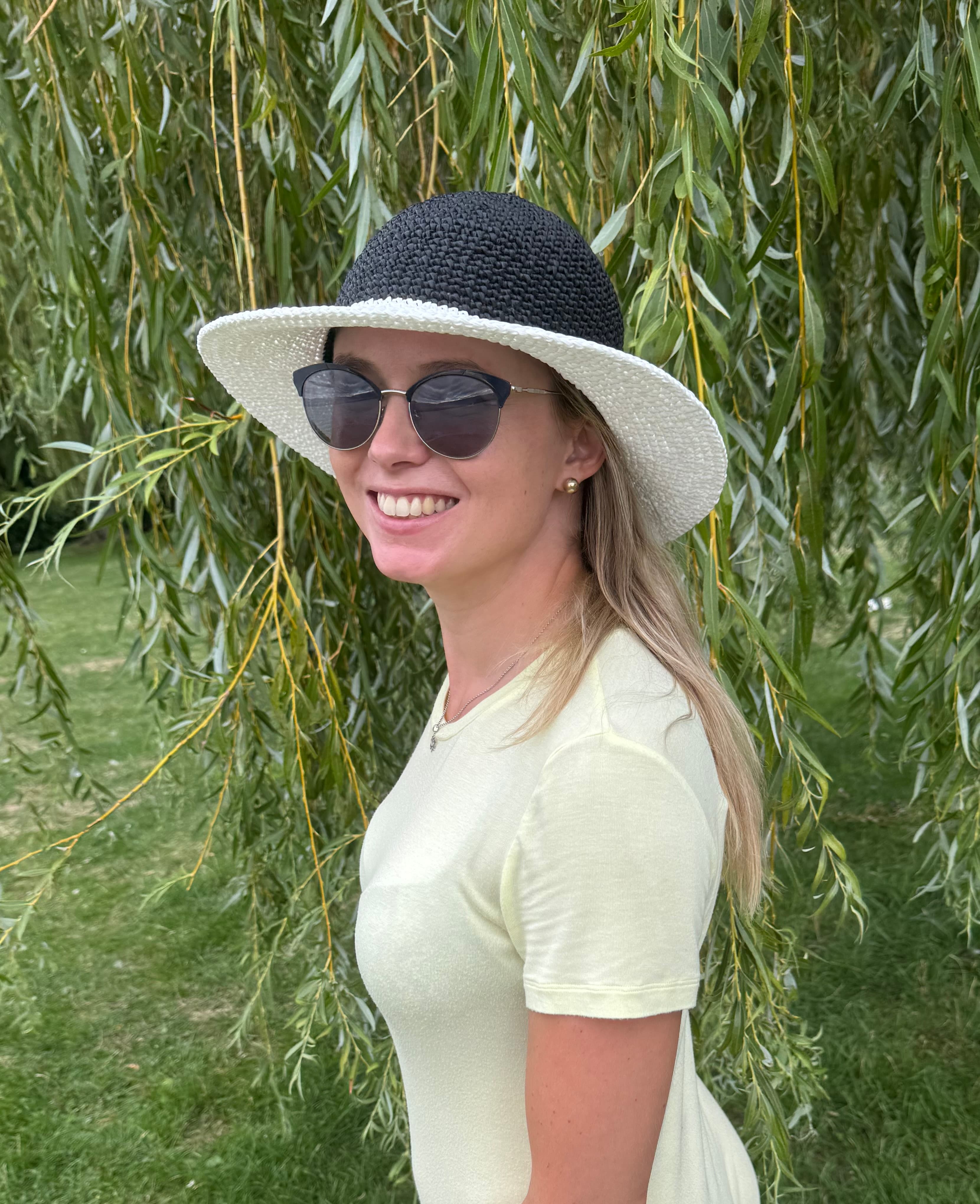 Femme souriante portant un chapeau noir et blanc et des lunettes devant un saule pleureur.