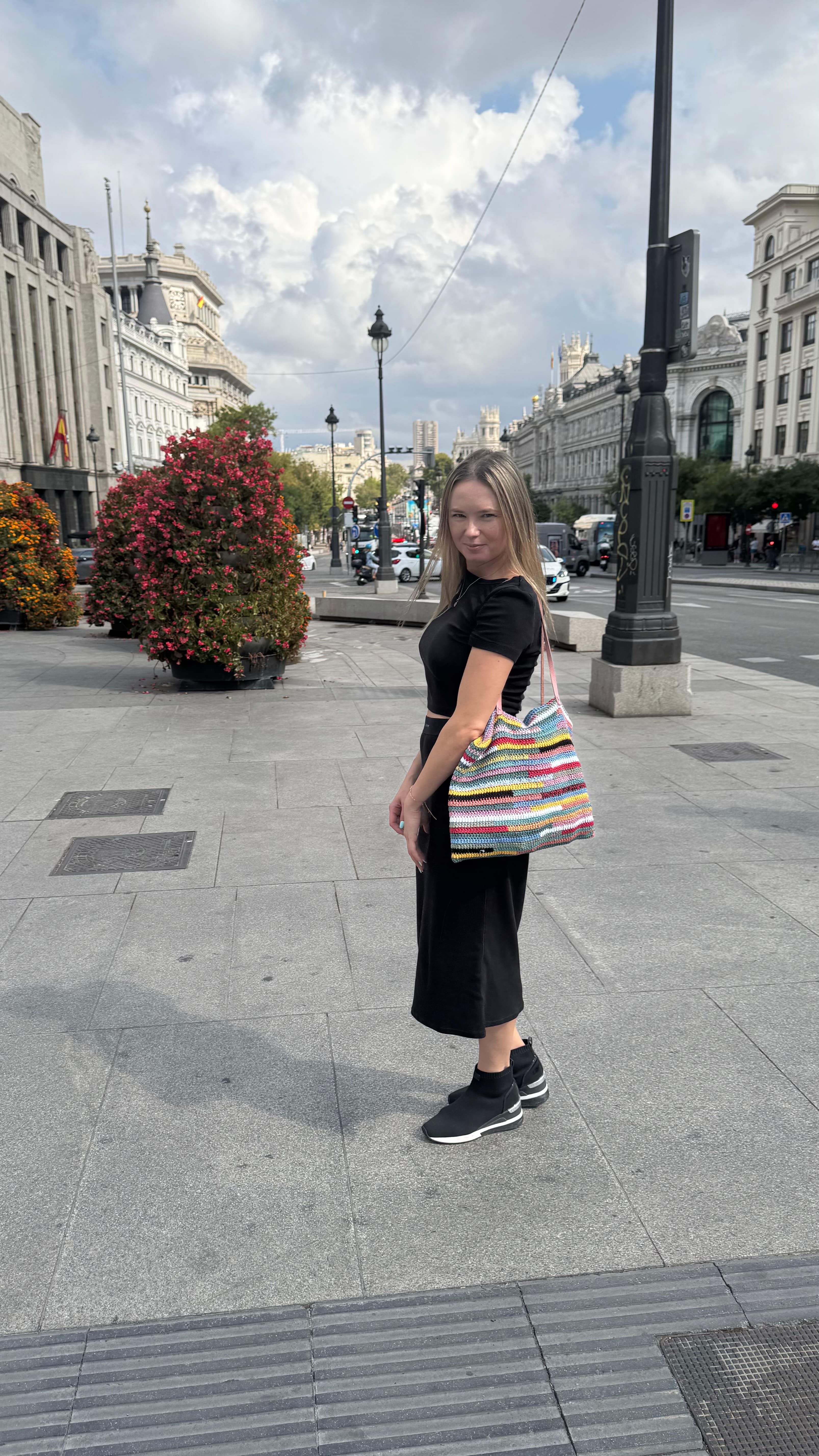 Femme en tenue noire avec un sac rayé multicolore sur un trottoir de ville.