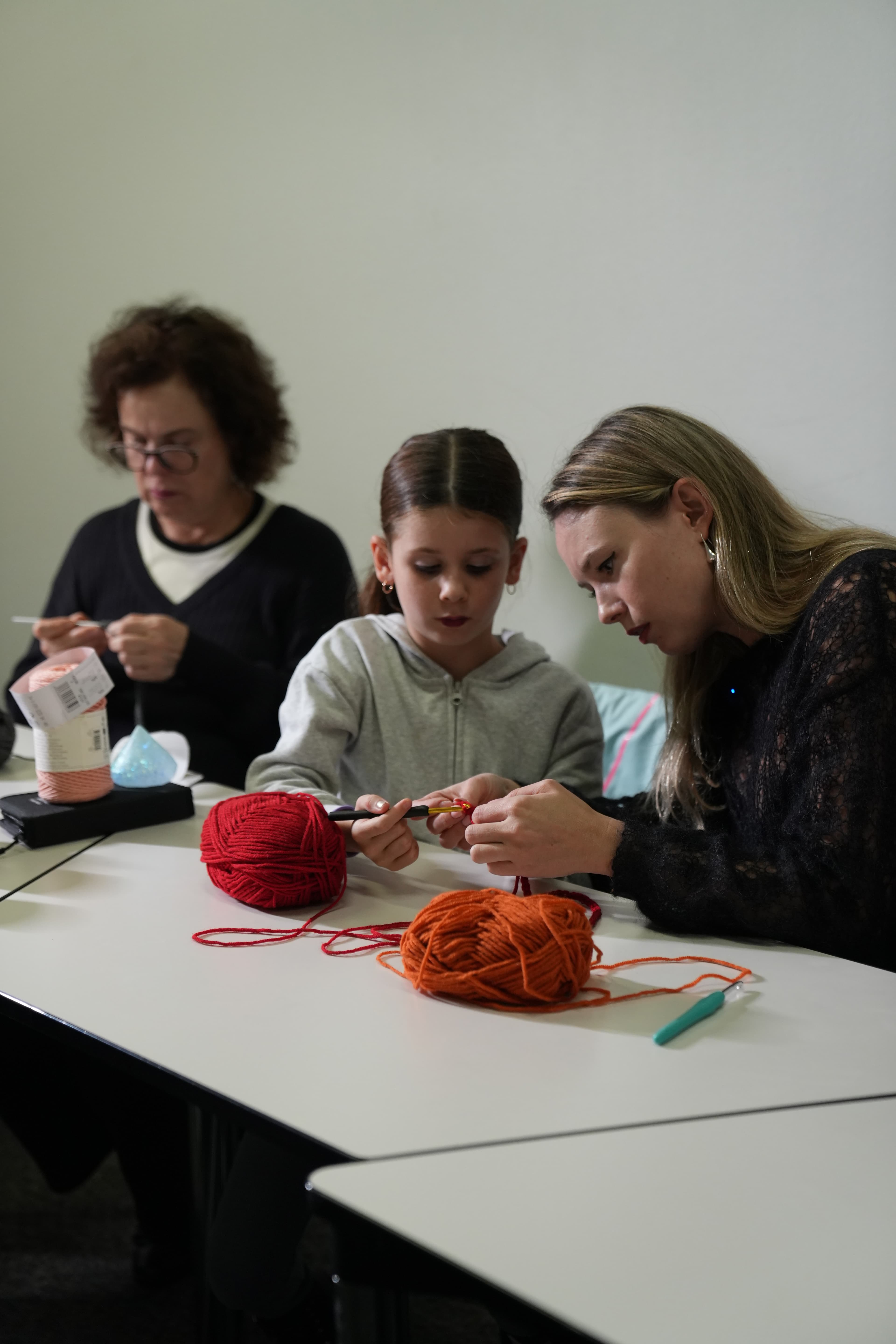 Une femme aide une petite fille à crocheter avec de la laine rouge et orange.